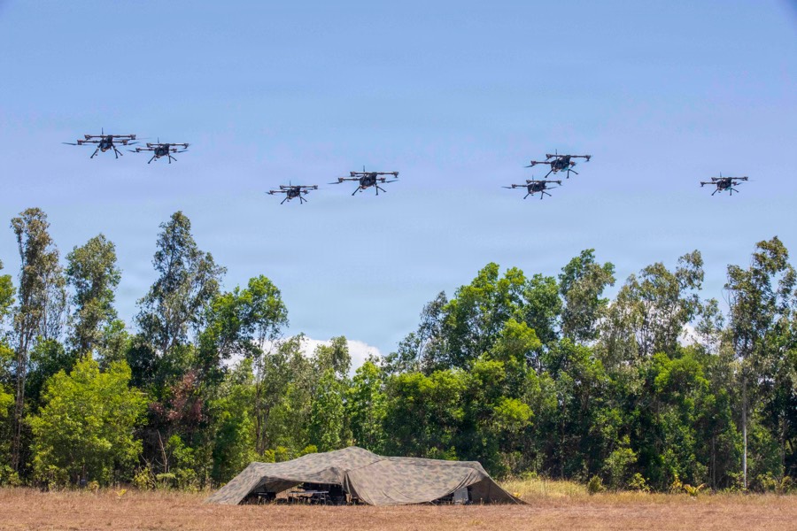 A swarm of small drones in flight above a treeline, with a camouflaged shelter on the ground below.