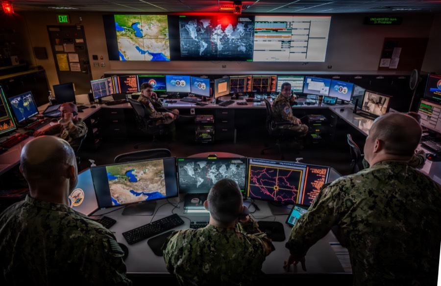 Service members at workstations facing a wall of large screens showing maps and data feeds.